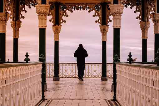 Color image depicting the rear view of a man, in silhouette against the sunset, standing alone surrounded by the Regency style architecture of a bandstand on the seafront in Brighton, England.