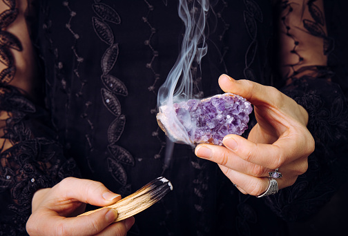 Close up view of woman in black lace dress, cleansing amethyst crystal cluster gemstone by smudging Palo Santo wood stick. Remove negative energy.