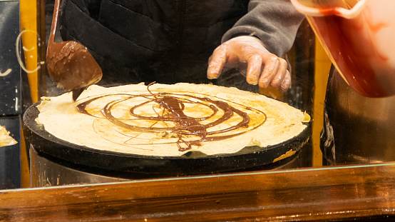 Stock photo showing unrecognisable person making crepes on electric cooker hob at street food market stall. The food vendor, who is wearing transparent disposable plastic gloves for food hygiene is spreading chocolate sauce over a cooked pancake.