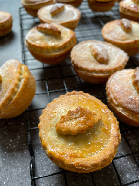 Photo showing a close-up view of festive dessert of deep filled mince pies on a metal, wire cooling rack. The traditional, individual tarts are topped with holly leaf shaped pastry and sprinkled with icing sugar are made with crispy, shortcrust sweet pastry and a sweet mincemeat filling. lote de imágenes de primer plano de pasteles de carne picada caseros, tradicionales y rellenos profundos espolvoreados con azúcar glas, postre navideño festivo, pastelería crujiente con carne picada dulce (mezcla de frutas secas picadas, licores destil