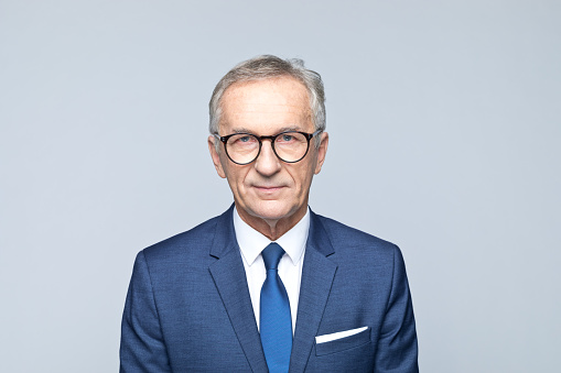 Portrait of handsome senior man wearing navy blue suit, white shirt and tie, looking at camera. Studio shot of male entrepreneur against grey background.