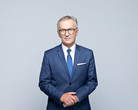 Portrait of handsome senior man wearing navy blue suit, white shirt and tie, looking at camera. Studio shot of male entrepreneur against grey background.