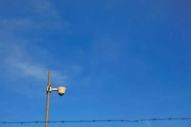 Surveillance Camera.Video surveillance.Camera behind the barbed wire on bright blue sky background.Regime object