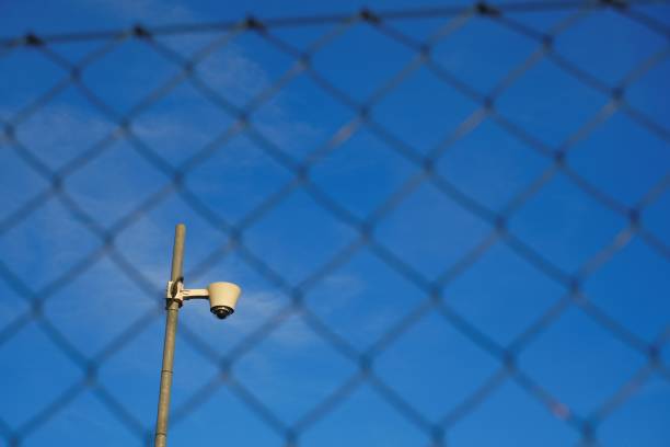 istock Surveillance Camera.Video surveillance concept. Camera behind metal grid on bright blue sky background. Closed area. 1354029884