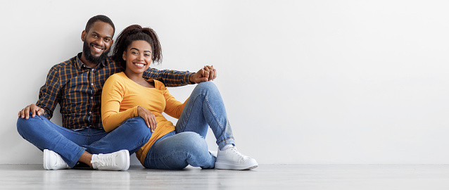 Smiling calm millennial african american couple, owners new apartment sit on floor on gray wall background, rest after moving, free space. Relocation and purchase of housing, rental of real estate