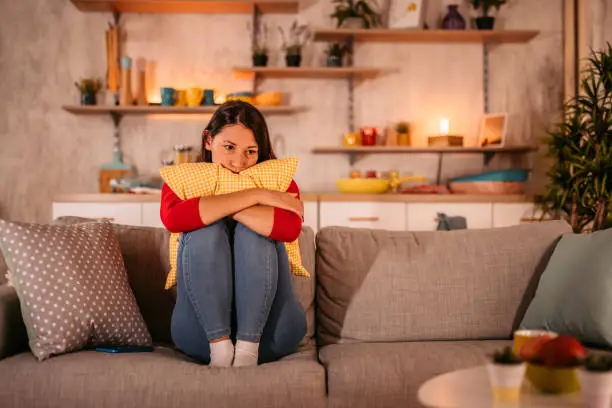 Woman looking depressed while sitting on sofa in living room and embracing pillow.