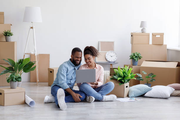 Glad young african american family resting, sitting on floor among cardboard boxes with things Glad young african american family resting, sitting on floor among cardboard boxes with things and plants in room interior and looking at laptop. Online shopping, furniture purchase design planning relocation stock pictures, royalty-free photos & images