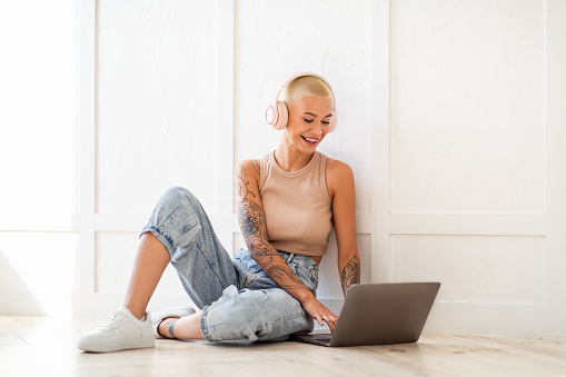 Young female freelancer in headphones working on laptop, sitting on floor and leaning on white wall. Woman having online job on computer, working remotely from home