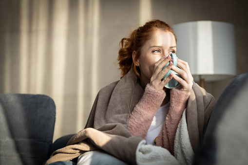 Woman with blanket over shoulders, siting on bed by the window in casual clothes enjoying her morning coffee