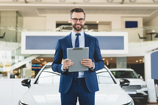 Successful confident smiling caucasian male shop assistant holding clipboard in formal clothes looking at camera at automobile car dealer shop