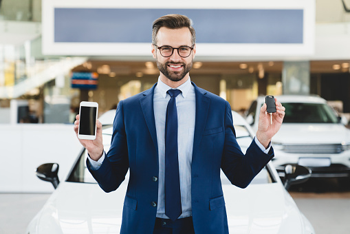 Successful caucasian businessman shop assistant in formal clothes standing at dealer shop holding new car keys with smart phone with mockup screen copyspace while buying selling new expensive car