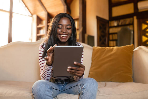 istock Happy young black lady using tablet, learning or working remotely, having online video call, sitting on sofa indoors 1354036167