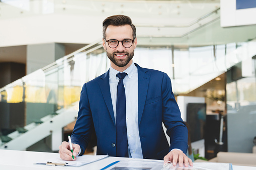 Successful caucasian smiling man shop assistant receptionist in formal attire writing looking at camera while standing at reception desk in hotel car dealer shop