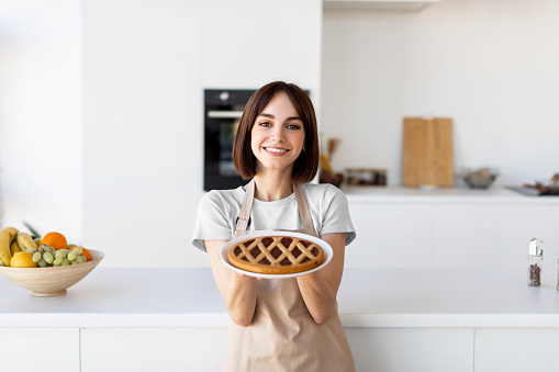 Homemade dessert. Happy lady holding and showing freshly baked pie, wearing apron and preparing pastry in modern kitchen interior, looking and smiling to camera