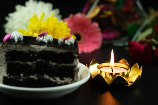 Golden Tealight Candle Illuminated In Lotus Shaped Holder With Slice Of Chocolate Truffle Cake On Side And Colorful Fresh Flowers On Dark Black Background