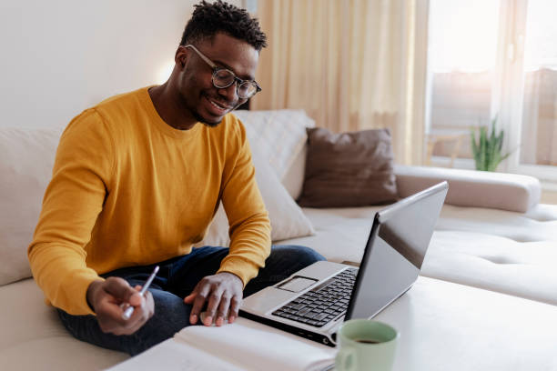 Handsome young businessman working on laptop computer at home. Cropped shot of a handsome young businessman sitting alone in his home office and working on laptop computer at home. Smiling black man using laptop in living room. Businessman using laptop at home. black man laptop stock pictures, royalty-free photos & images