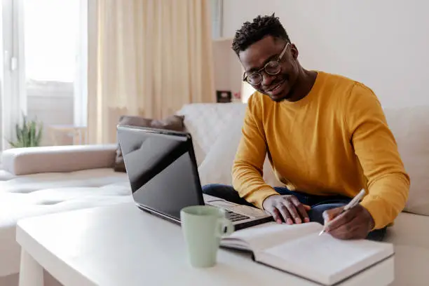 African young man in eyeglasses sitting at the table in front of laptop computer and looking at monitor at home, drinking tea. Handsome young African man using laptop and smiling while sitting indoors