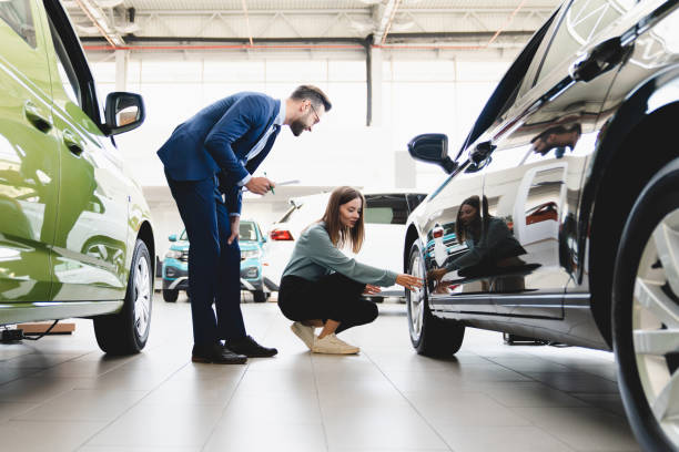 beautiful young caucasian female client customer choosing new car, trying checking its options, tire, wheels while male shop assistant helping her to choose it at dealer auto shop - 汽車經銷商陳列室 個照片及圖片檔