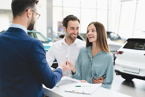 Buying purchasing new car. Male shop assistant congratulating young family couple with buying auto giving car keys at dealer shop.
