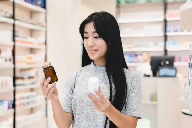 Photo of Female asian customer client looking for medications, medicines, comparing two jars with drugs, pills, vitamins, antibiotics, painkillers, choosing between two products in pharmacy drugstore