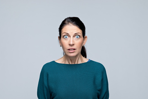 Portrait of shocked mature woman looking at camera with mouth open, rolling eyes. Studio shot of female entrepreneur against grey background.