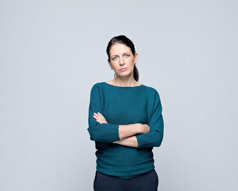 Portrait of sad mature woman standing with arms crossed and looking at camera. Studio shot of female entrepreneur against grey background.