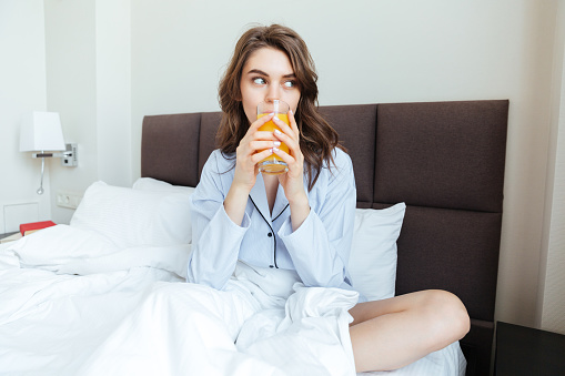 Portrait of a young pretty woman wearing nightwear and drinking orange juice while sitting on bed at home in the morning