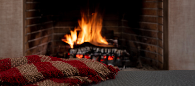 Relaxation near the fire, Christmas holiday cozy warm home interior. Blanket on the sofa, fireplace burning wood logs background.