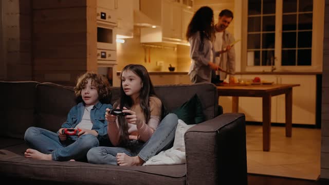 Children are playing on game console, parents preparing dinner at the background