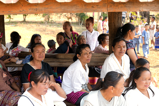 Group of sitting thai vilage women captured in center of rural small town in Chiang Mai province. They are sitting in shadow under a roof. In right background are many children