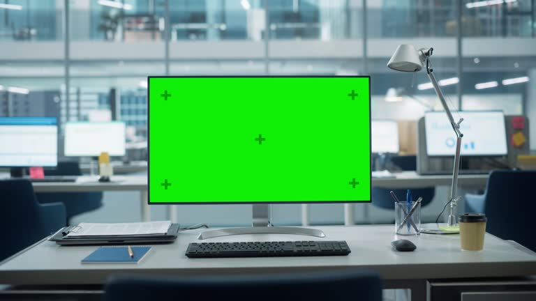 Desktop Computer Monitor with Mock Up Green Screen Chroma Key Display Standing on the Desk in the Modern Business Office. In the Background Glass Wall with Big City Office. Zoom Out Shot.