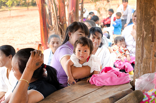 Larger group of rural thai women and one with a baby girl in small town and village in Chiang Mai province. Women are sitting together, some under a roof in shadow, In background are children and more people