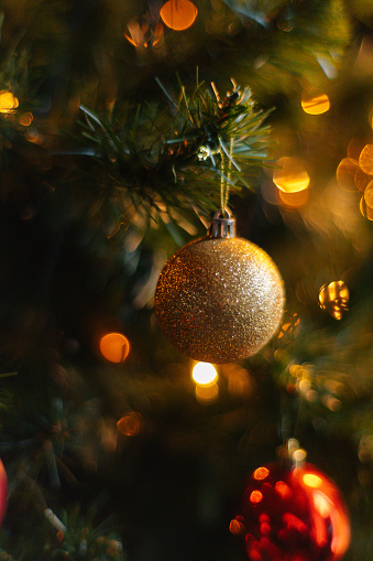 Close-up vertical shot of beautiful Christmas tree decorated with festive golden ball and warm lights at home, selective focus. Holiday New Year tree with colorful baubles and illuminated garland.