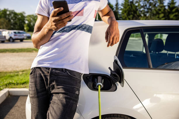 istock Man using mobile phone while charging electric car outdoors 1354070048
