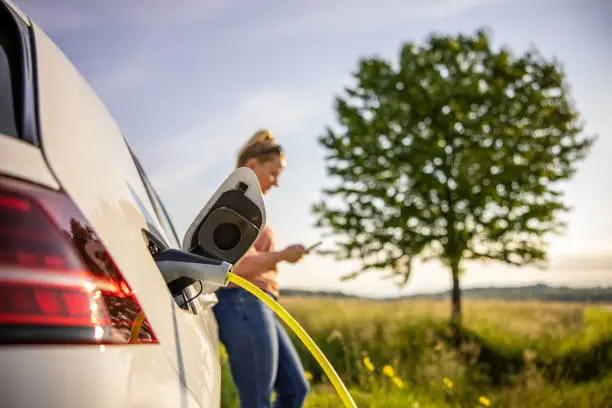 Woman text messaging using mobile phone while charging electric car on green landscape