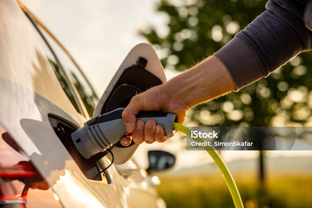 Man inserts a power cord into an electric car for charging in the nature Hand of man inserting a power cord into an electric car for charging ecofriendly vehicle on green landscape Electric Car Stock Photo