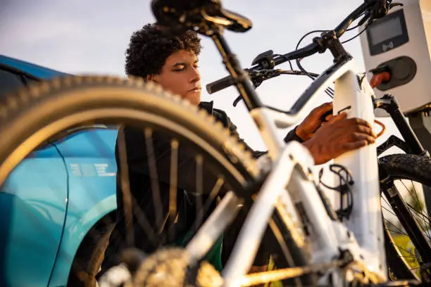 Young man inserting plug into electric bicycle for charging at electric charging station