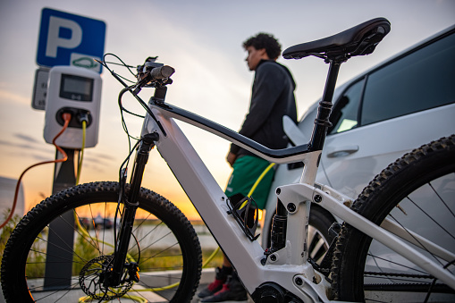 Young man leaning on car while his electric bicycle and electric car being charged at the electric vehicle charging station