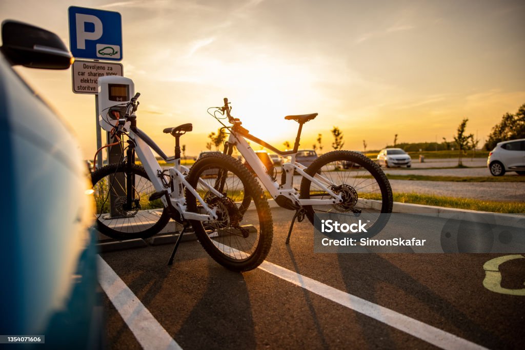 Two electric bicycles being charged at the electric vehicle charging station Two electric bicycles being charged at the electric vehicle charging station on parking lot during sunset Electric Bicycle Stock Photo