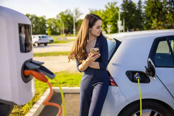 Young woman in dress holding mobile phone while waiting for electric car to charge at charging station