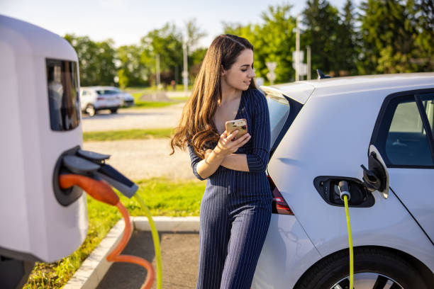 young woman holding mobile phone while waiting for electric car to charge - electric car imagens e fotografias de stock