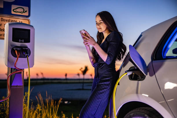 istock Young woman text messaging on mobile phone while waiting for electric car to charge 1354072001