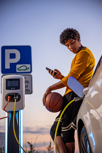 African american basketball player using mobile phone while he is waiting for his electric car to be charged at charging station