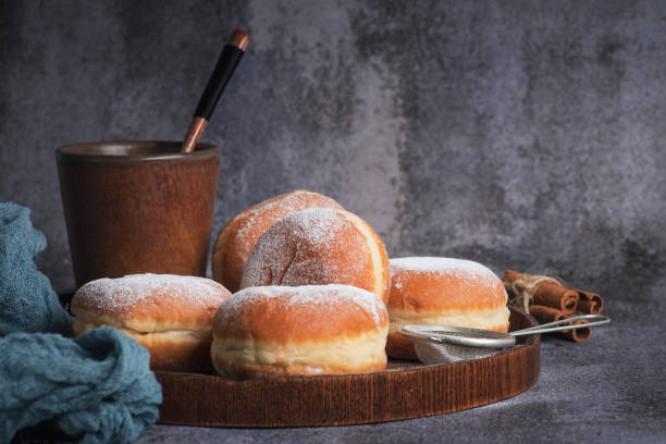 A few donuts filled with jam sprinkled with powdered sugar in a black bowl with a cup of milk on rustic wooden background
