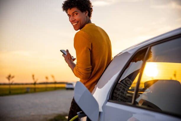 Man using mobile phone while charging electric car African american man using mobile phone while he is waiting for his electric car to be charged at charging station man looking at phone over shoulder stock pictures, royalty-free photos & images