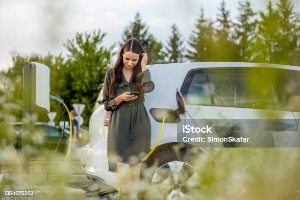 Frau Mit Handy Während Sie Auf Elektroauto Auf Dem Parkplatz Aufladen Stockfoto und mehr Bilder von Elektroauto