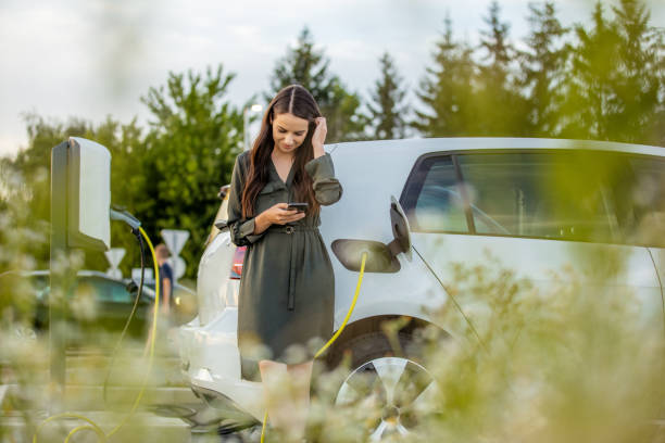 Woman using mobile phone while waiting for electric car to charge in the parking lot Front view three quarter length shot of a young woman in a dress using her mobile phone while waiting for her white electric car to charge tesla motors stock pictures, royalty-free photos & images