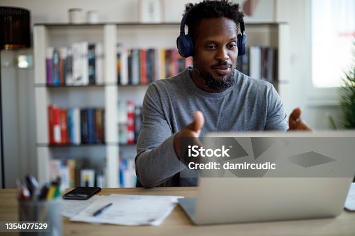 istock Man with bluetooth headphones having video call on laptop computer in his home office 1354077529