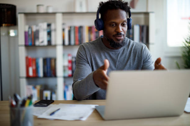 homem com fones de ouvido bluetooth tendo chamada de vídeo em computador portátil em seu escritório - working man audio - fotografias e filmes do acervo