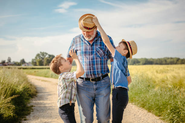 Grandsons pulling grandfather hat while walking on dirt road beside field